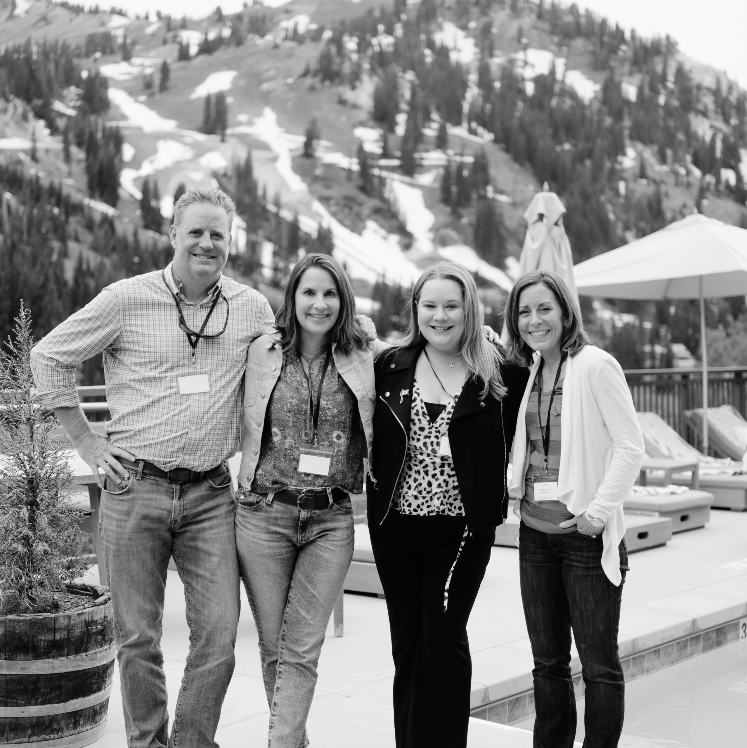 Black and white photo of for business people, three women and one man, posing and smiling outside on a deck for photo, with mountains and pool in the background.
